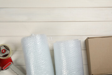Bubble wrap rolls, cardboard box and tape dispenser on white wooden background, flat lay. Space for text