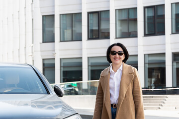 A beautiful confident woman is standing near a car on the background of a business center and smiling.