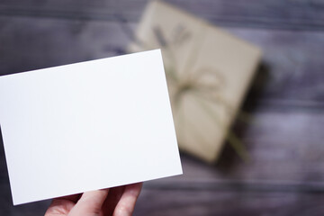 Woman's hand holding a blank white card. Surprise gift mock-up scene. Selective focus. Present on wooden background. Empty space for text. Copy space.