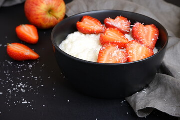 Delicious rice porridge in milk with strawberry pieces and coconut flakes. On a black background. Cooking.