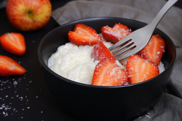 A fork pricks the strawberries. Delicious rice porridge in milk with strawberry pieces and coconut flakes. On a black background. Cooking.