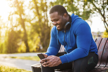 Man with headphones using mobile phone while resting after exercise on bench.
