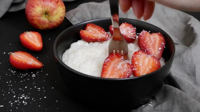The girl pricks a piece of strawberry on a fork. Delicious rice porridge in milk with strawberry pieces and coconut flakes. On a black background. Cooking.