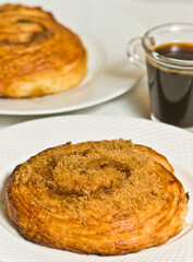 Top, front view, close distance of a freshly baked, croissant cinnamon role on a white, round plate, with a glass cup of espresso, in a red and white stripped place mat