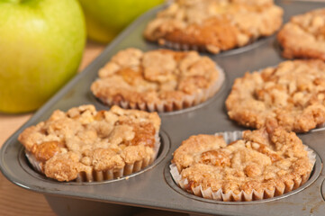 top, front view, close up of freshly baked apple crumb muffins in paper molds and baking pan, cooling with two green apples in background