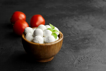 Mini mozzarella cheese balls with ripe tomatoes in wooden bowl on black stony background