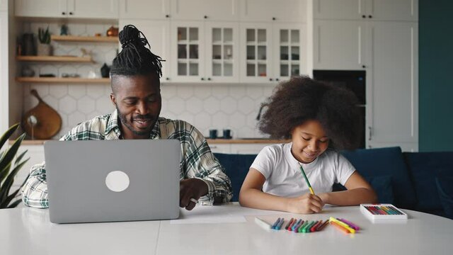 Young african american father working on laptop computer, his little daughter drawing with crayons near him at home