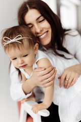 Happy brunette woman gently hugging beautiful baby girl, smiling. Attractive young mother holding little daughter in arms, maternity concept.