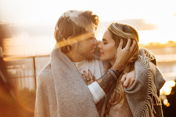 Portrait of happy beautiful couple at the sunset, cuddling in cozy blanket. Charming loving blond man hugging attractive woman, enjoying tenderness, spend time together, romantic date concept - obrazy, fototapety, plakaty