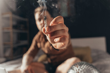 Man offering a hand rolled cannabis cigarette while sitting at the bed - obrazy, fototapety, plakaty