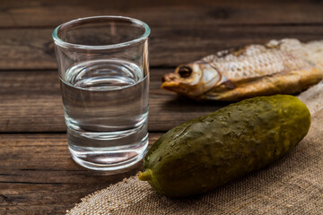 Shot of vodka with piece of cloth and wet pickled cucumber with a stockfish on an old wooden table. Close up view, focus on the cucumber