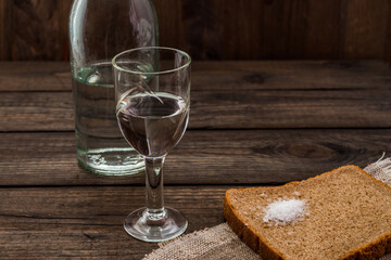 Shot of vodka with bottle and a piece of black bread with salt and cloth cut on an old wooden table. Angle view