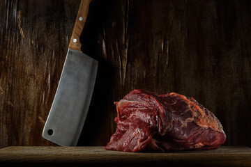 piece of fresh raw beef on a cutting board with a kitchen cleaver near an old wooden wall. artistic moody photo in rustic style with copy space