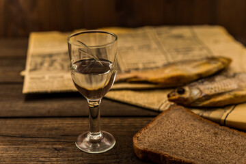 Glass of vodka with a stockfish and newspaper with piece of the black bread on an old wooden table. Angle view