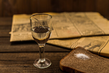 Obraz premium Glass of vodka and newspaper with piece of the black bread on an old wooden table. Angle view, shallow depth of field