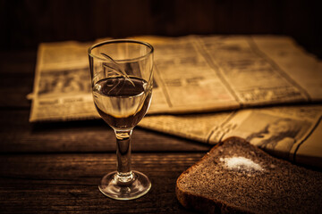 Glass of vodka and newspaper with piece of the black bread on an old wooden table. Angle view, shallow depth of field