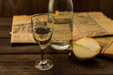 Glass of vodka with  newspaper and onion with bottle on an old wooden table. Angle view, shallow depth of field, focus on the glass of vodka - obrazy, fototapety, plakaty