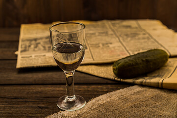 Glass of vodka with  newspaper and pickled cucumber with canvas on an old wooden table. Angle view, shallow depth of field, focus on the glass of vodka