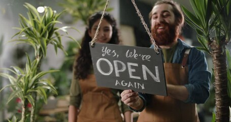 Positive employees reopening flower shop after COVID-19 pandemic. Happy greenhouse workers hanging open again sign at door - small business people after quarantine 4k footage