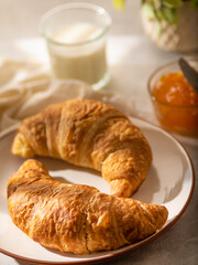 Croissants on a white plate. Nearby is orange jam and milk in a glass. Beige paints. White background. Pastel shades. close-up.