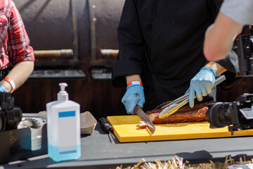Chef slicing delicious bbq meat at street market on table with sanitizer. Vendor in gloves cooking tasty smoked steaks at street food festival, making video. Summer vacation picnic