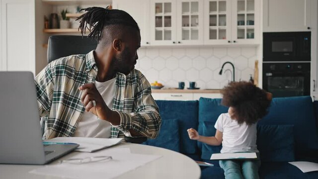 African american man video calling to partners, talking to drawing daughter on couch and keep talking, working at home