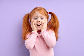 surprised girl with natural red hair and freckles, stands over purple background touching cheeks, with wide opened mouth, look at camera in shock