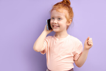Redhead child girl communicates on mobile phone, smiles and talks isolated on purple background. Closeup smiling kid talking on smartphone. People sincere emotions lifestyle concept