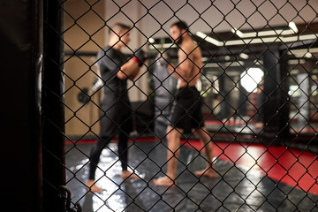 Two strong men boxers boxing, exercising thai boxing in silhouette, MMA. View through cage, in ring at gym. Side view. Focus on cage, close-up