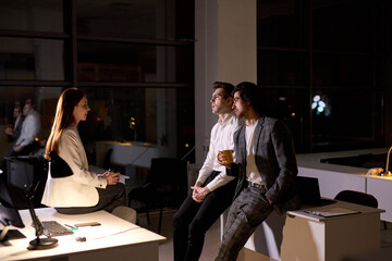 Colleagues in formal outfit stand having conversation, taking a break at late night in boardroom, smiling, discussing, sharing ideas, explaining plans. teamworking, office workers concept