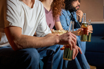 friends supporting football team at home, cheering for favourite team, feel concern. sit on sofa at home, in dark room at night, drinking beer, focus on hands with bottle. side view