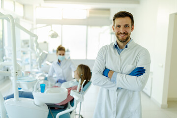 Male dentist standing with his hands crossed, wearing gloves and white coat and looking at camera.