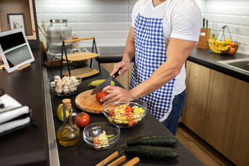 Muscular chef, wearing apron, slicing a tomato in the kitchen of his apartment