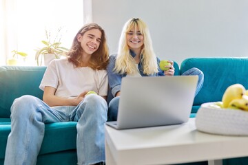 Laughing couple of teenagers having fun, sitting on couch, looking at laptop