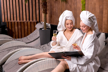 young women in spa salon in towel and bathrobe looking at prices of procedures drinking champagne,...