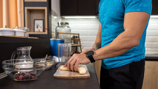 Close up of a male athlete in the kitchen slicing a banana while preparing a shake