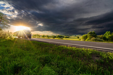 Blue truck driving on the asphalt road in rural landscape in the rays of the sunset with dark storm cloud