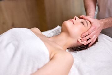 Side view on relaxed woman getting face massage in beauty spa.Caucasian female with closed eyes relaxing in spa while getting head massage. Serene calm woman relaxing indoors in beauty center.