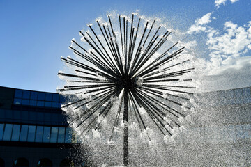 A round spherical fountain that spews water under pressure. The globular structure is similar to a dandelion. Water bris. Windows of a modern building in the background.