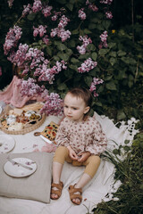 little girl with a serious face sits on a white picnic blanket. a gloomy girl is waiting for guests at her picnic. children's picnic in the park or garden.