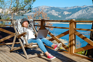 Young woman at the deck of lakehouse, sitting on a wooden chair. Female freelancer working on her laptop sitting outside of the lodge with beautiful lake view. Copy space, close up, background.