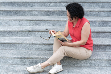 A woman sits on the steps in the city and reads a book.