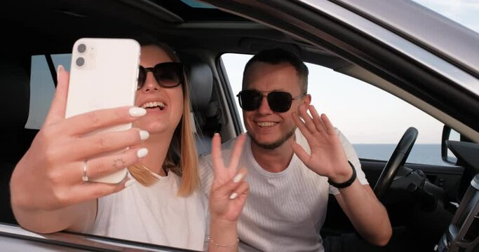Young Man and Woman Sitting in Car with Sea View from Window and Using Smartphone for Video Call, Waving Hands to Say Hello During Their Road Trip