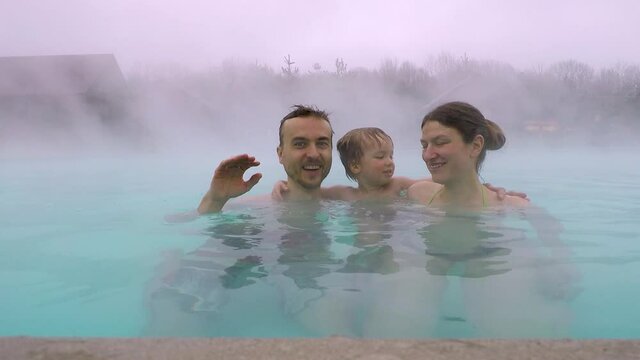 Family having fun on geothermal spa resort. Father, mother and son relaxing in outdoor hot spring pool in mountains when weather is cold and rainy. Making content for travel vlog.