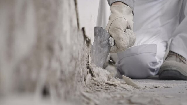 Construction worker hands with gloves working with spatula scrape off the plaster from the wall for house renovation, close up with rubble and tools