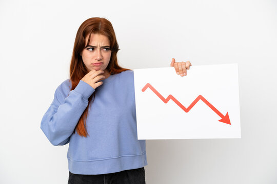 Young redhead woman isolated on white background holding a sign with a decreasing statistics arrow symbol and thinking
