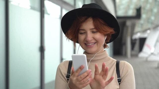 Smiling red haired woman wearing stylish hat using mobile phone communication online, walking on the street. Travel concept, slow motion