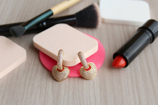 Par de brincos de ouro com brilhantes, cristais, diamantes no formato de cora&ccedil;&atilde;o. Fundo de madeira com esponjas, batom e pinceis de maquiagem.