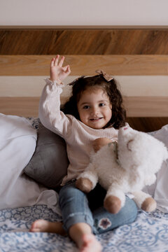 Hispanic toddler girl lounging in bed with her favorite stuffed animal-Little girl smiling as she raises her hand
