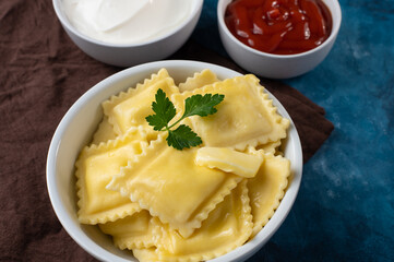 In the photo, ravioli in a beautiful earthenware white bowl with a sprig of mint, as well as sour cream and tomato sauces are also in a white dish. Beautiful blue background. No people.